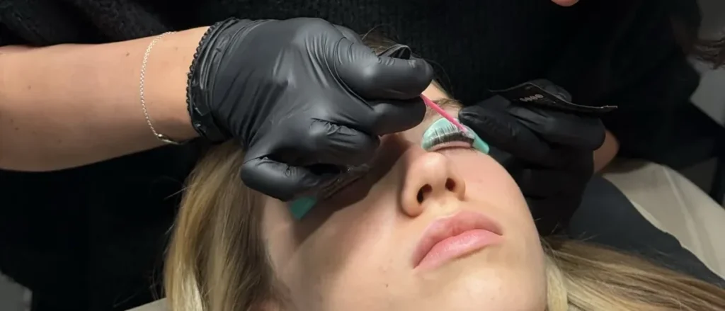 Behandschuhte Hände bearbeiten die Wimpern auf einem Silikonpad am geschlossenen Auge.