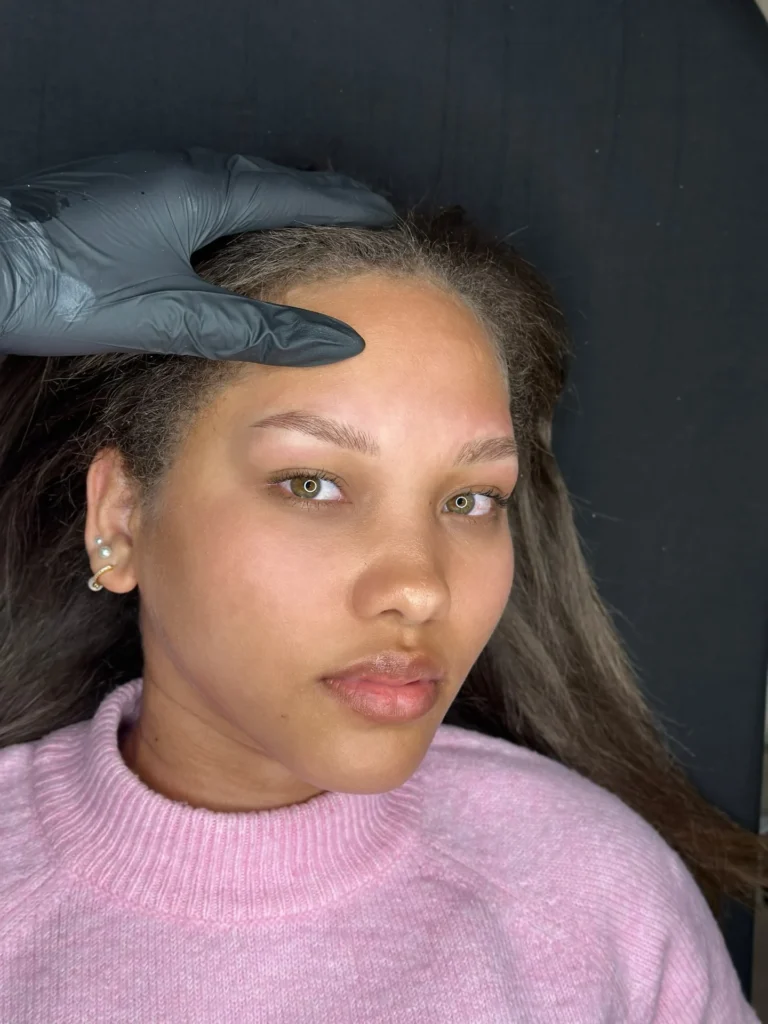 Gesicht mit feinen Augenbrauenhaaren und sichtbarer Kontur; eine behandschuhte Hand hält das Haar zurück.
