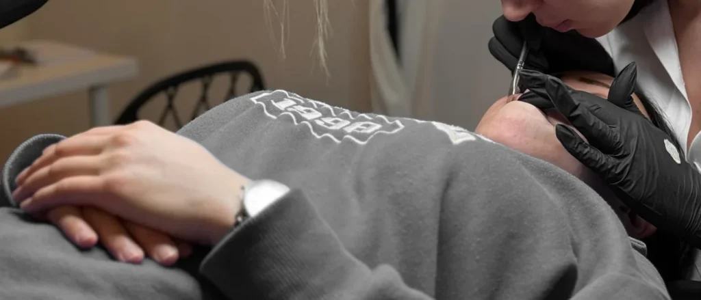 Behandschuhte Hände arbeiten mit einem Handstück im Augenbrauenbereich.